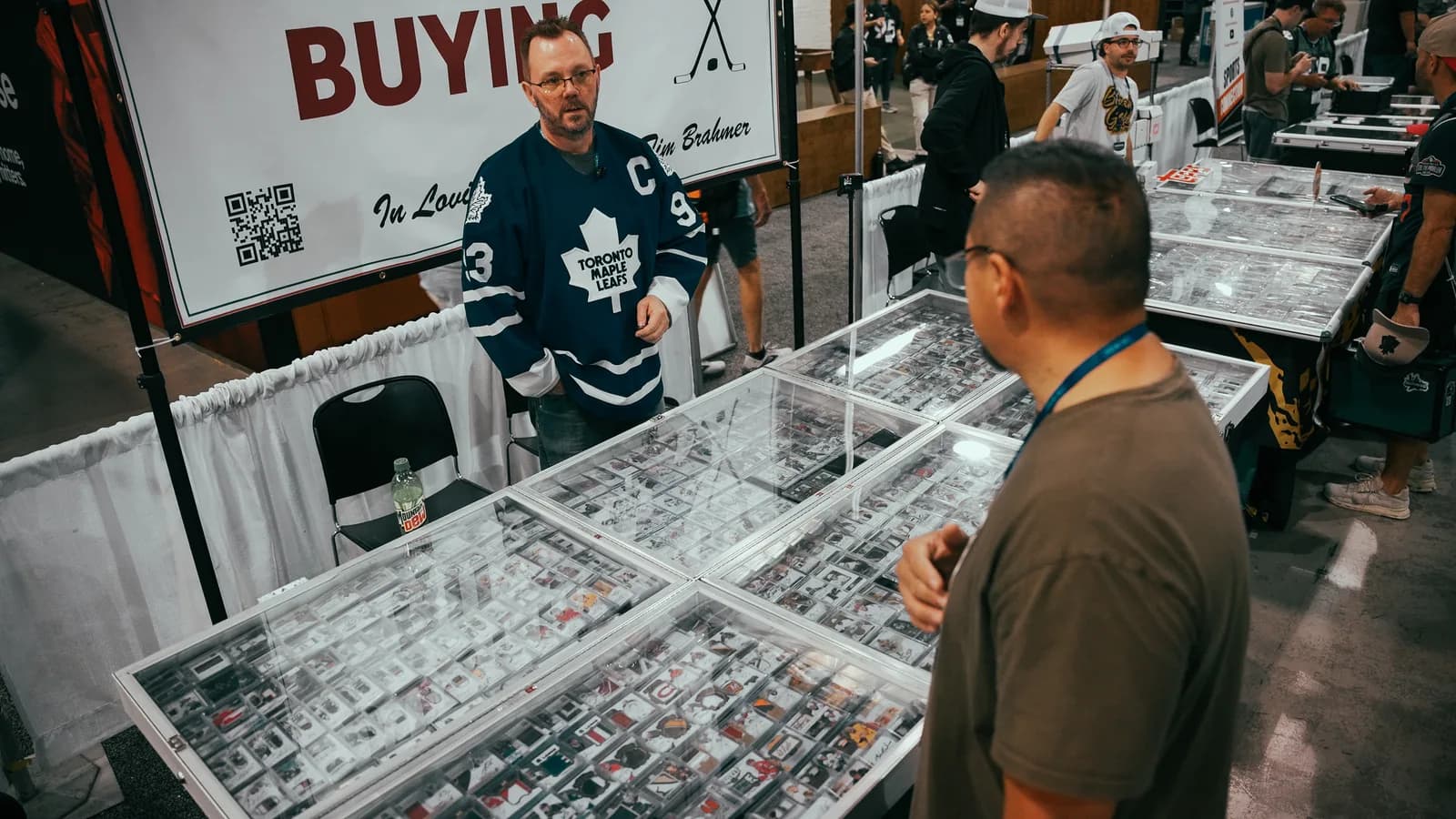 A dealer in a Toronto Maple Leafs jersey standing behind glass cases of graded cards beneath a hand-lettered BUYING sign, one collector leaning in to study the rows