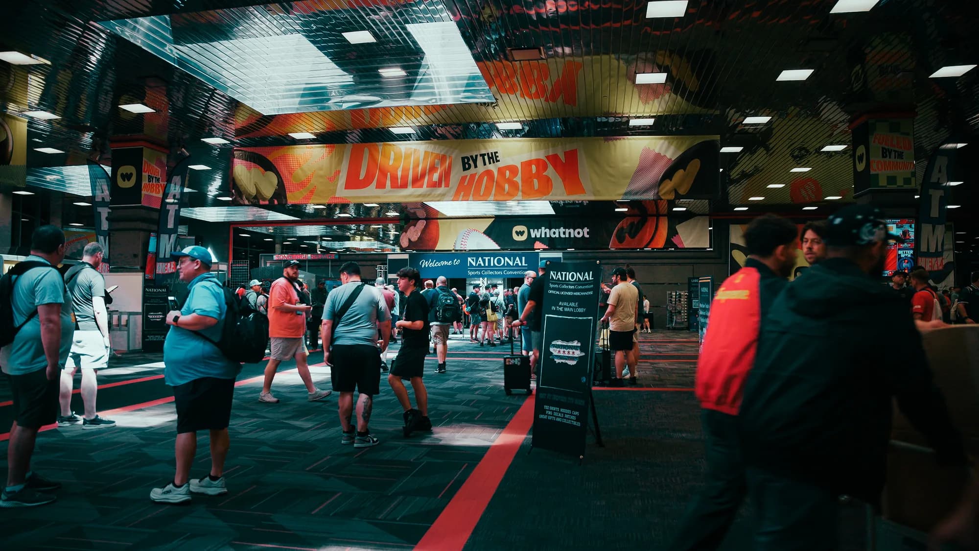 Attendees walking in beneath a large DRIVEN BY THE HOBBY banner and a Whatnot sign at the main entrance to the National Sports Collectors Convention, past the Welcome to National archway