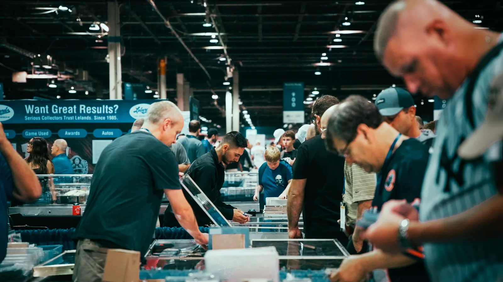 Collectors bent over glass showcases along a dealer row at The National, hands in the cases, a Lelands banner reading “Serious Collectors Trust Lelands. Since 1985” behind them
