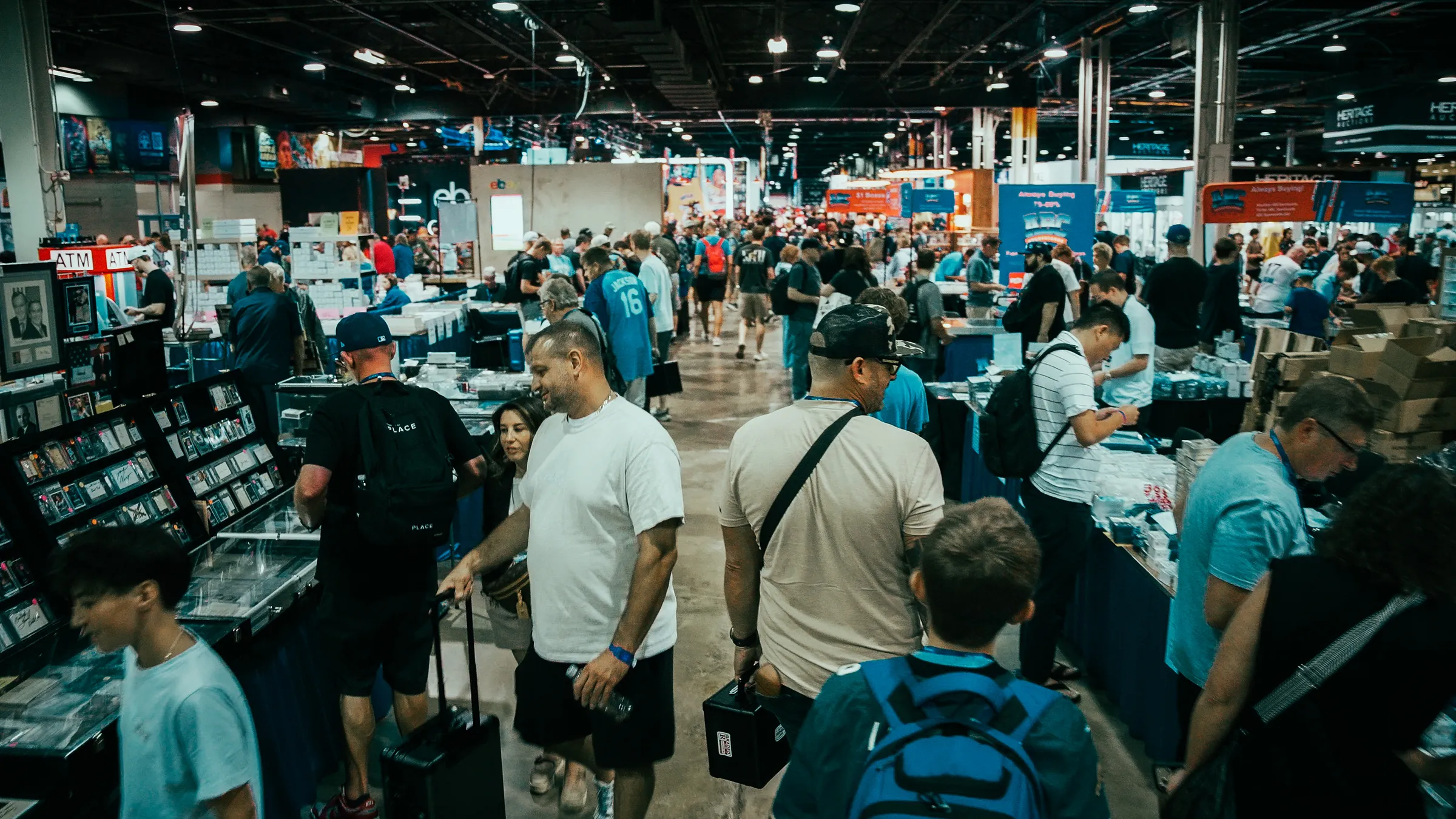 A packed main aisle at the 2026 National in Rosemont, Illinois: collectors moving between dealer tables with cases and carry-ons, the eBay and Heritage Auctions booths visible down the hall